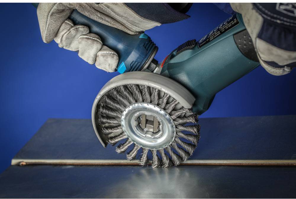 Eine Person hält ein Elektrowerkzeug mit Drahtbürste, poliert eine Metalloberfläche. Schutzhandschuhe sind getragen.