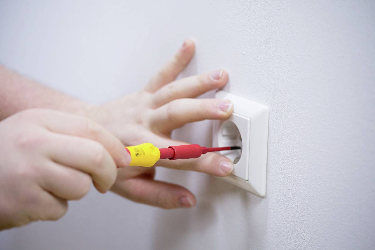 Eine Person repariert eine Steckdose mit einem Schraubenzieher an einer weißen Wand.
