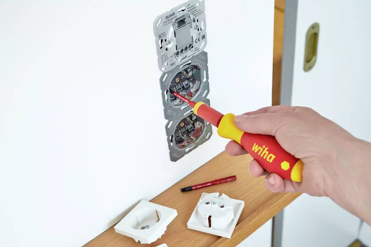 Eine Person ersetzt elektrische Steckdosen mit einem isolierten Schraubendreher, wobei Steckdosenabdeckungen und ein Werkzeug auf einer Holzoberfläche in der Nähe liegen.