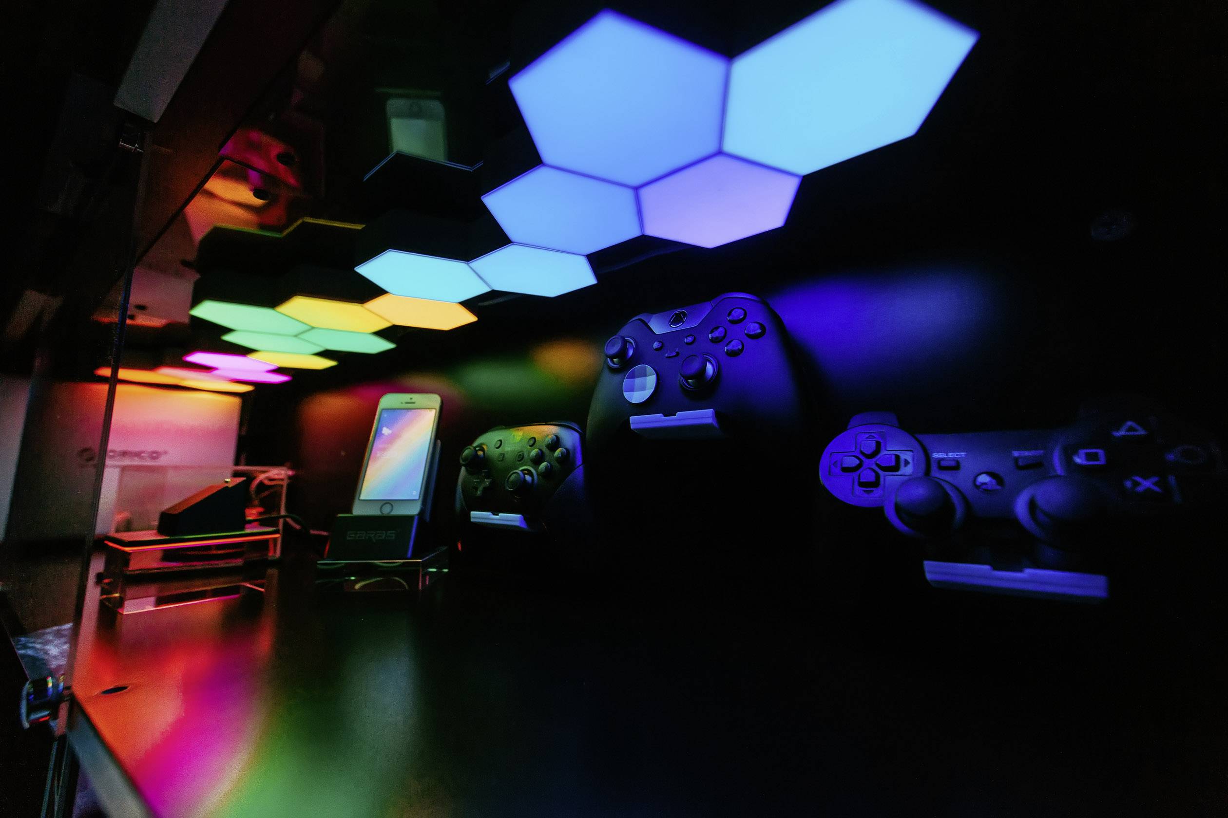 Gaming-Setup mit beleuchteten LED-Panels an der Wand. Zwei Controller und ein Smartphone befinden sich auf der Ablage darunter.