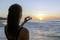 Eine Frau am Strand fotografiert einen Sonnenaufgang über dem Meer. Im Vordergrund ist das leuchtende Wasser und die klare Morgensonne sichtbar.