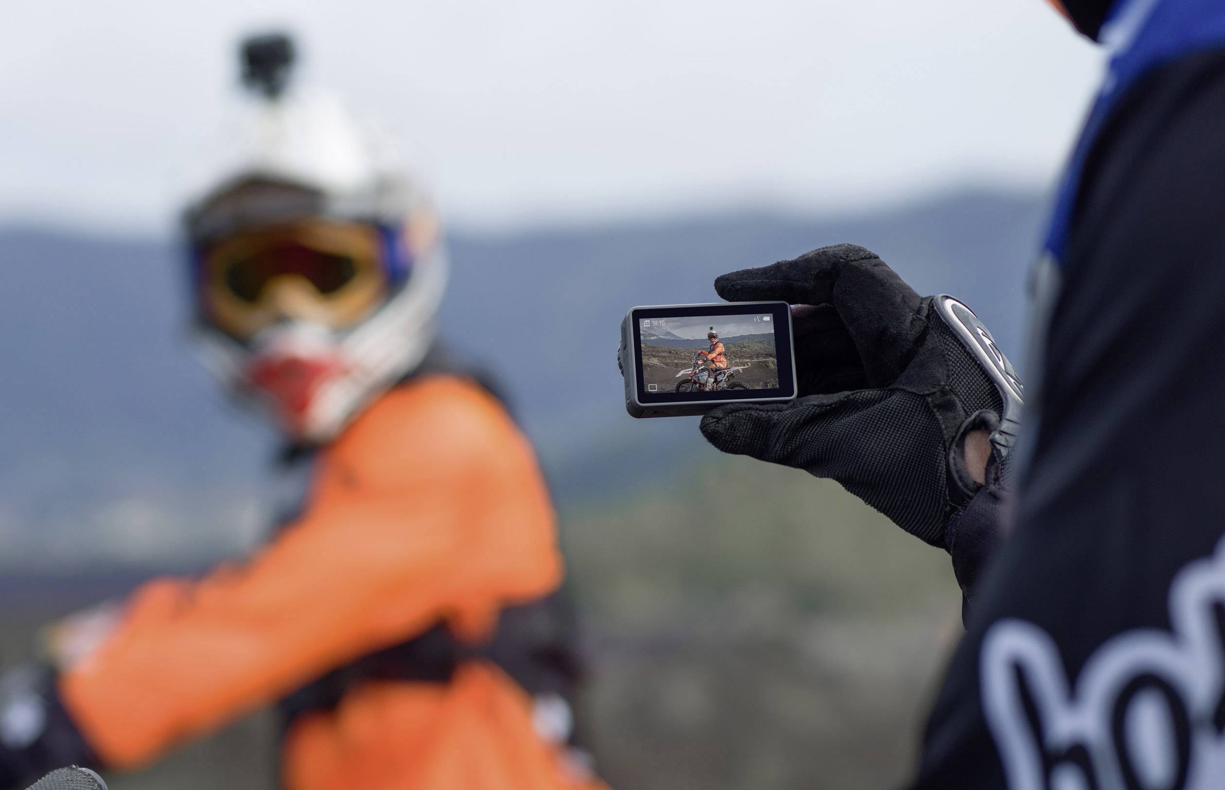 Eine Person in orangefarbener Motorradbekleidung wird aus der Ferne mit einer Action-Kamera gefilmt. Die Szene zeigt eine Landschaft im Hintergrund.
