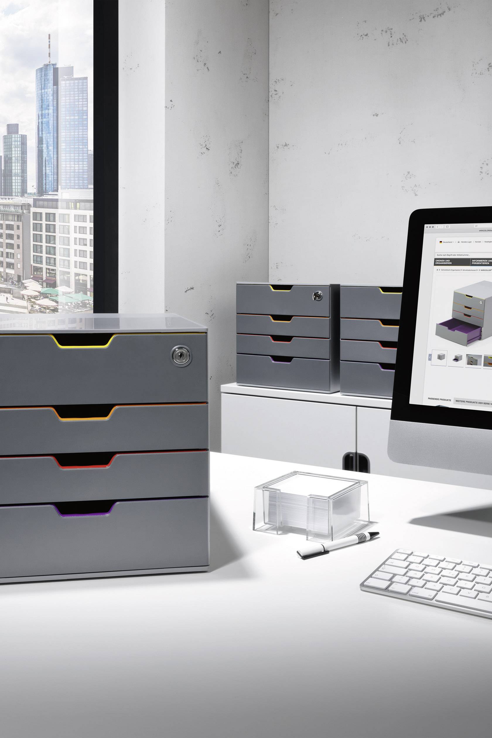 Ein modernes Büro mit Blick auf die Stadt. Auf einem Tisch stehen silberfarbene Schubladenboxen, ein Computer zeigt ein Produktbild.