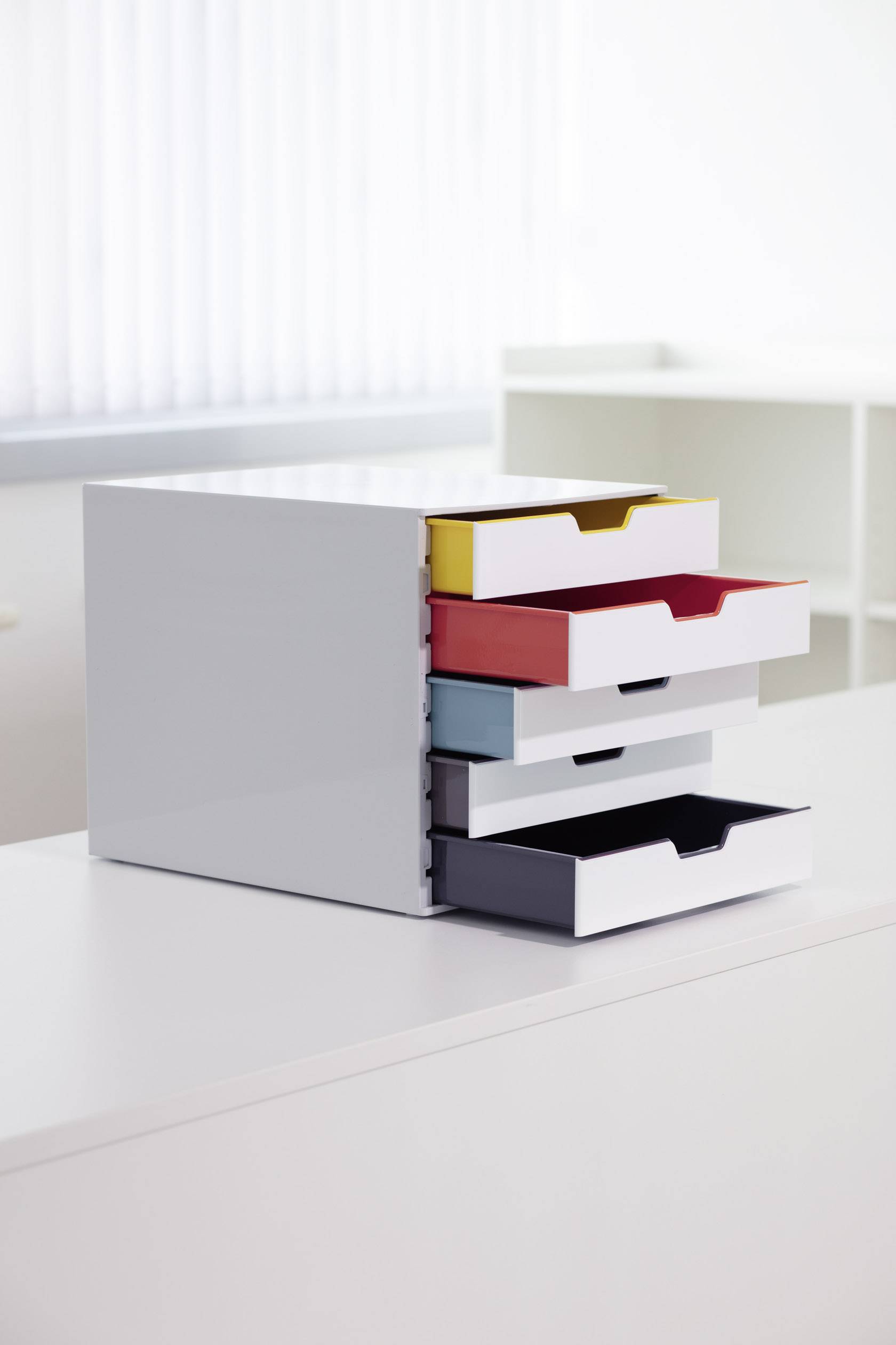 Kleiner Schubladencontainer mit vier farbigen Schubladen (gelb, rot, blau, schwarz) steht auf einem weißen Tisch in einem Büro.