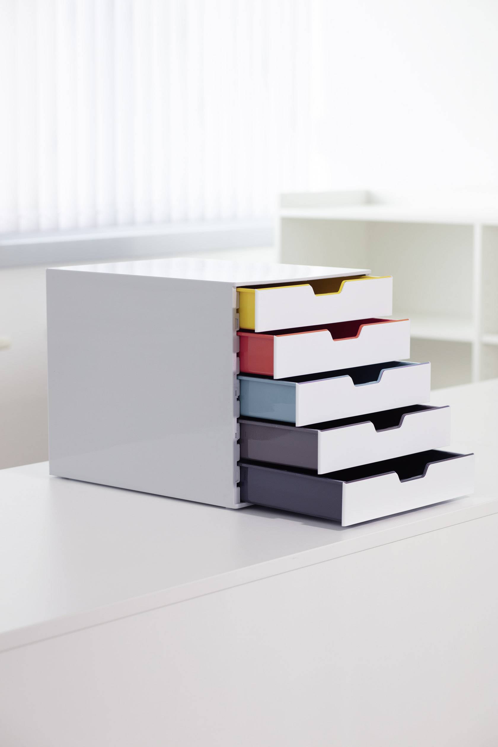 Fünfschubladen-Organizer enthält bunte, herausgezogene Schubladen für Bürobedarf. Steht auf einem weißen Tisch in einem hellen Raum.