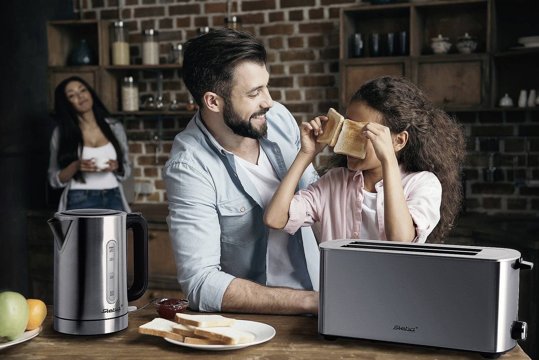 Ein Mann und ein Mädchen spielen mit Toast vor einem Toaster, während eine weitere Person im Hintergrund lächelt. Ein Wasserkocher steht daneben.