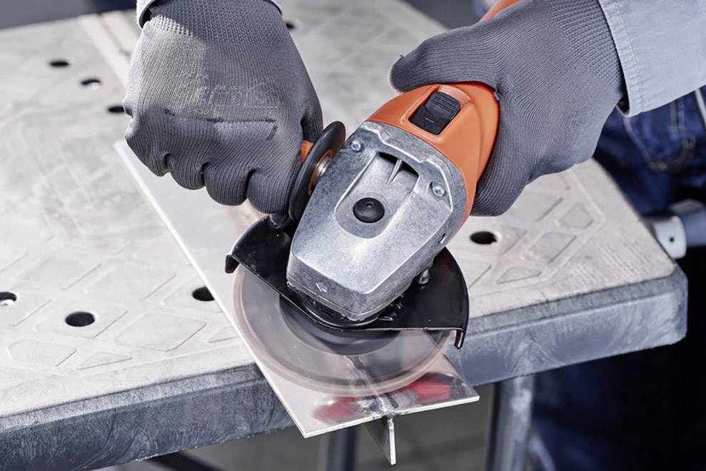 Eine Person schneidet mit einem Winkelschleifer ein Stück Metall auf einem Tisch. Schutzbekleidung wird getragen.