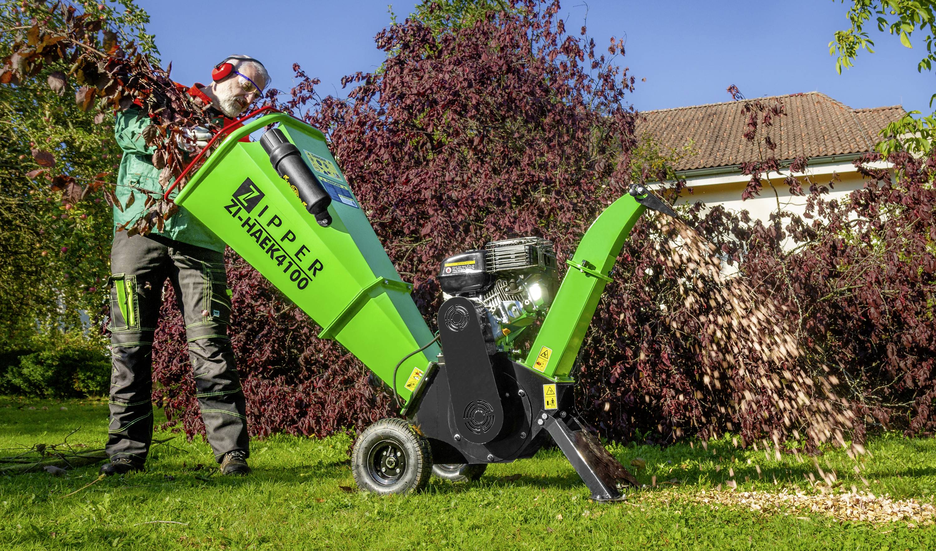 Eine Person bedient draußen einen grün-schwarzen Häcksler. Zweige werden eingeführt, während Holzspäne seitlich ausgeworfen werden.