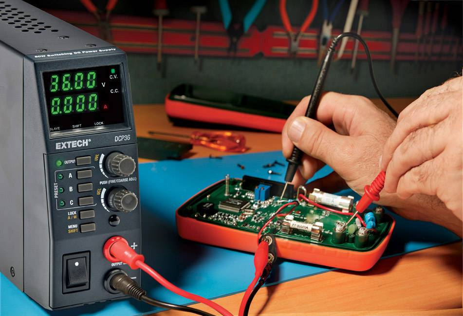 Eine Person testet eine elektronische Platine mit einem Multimeter. Ein Netzgerät zeigt '36.00 V' an. Technik-Arbeitsplatz.