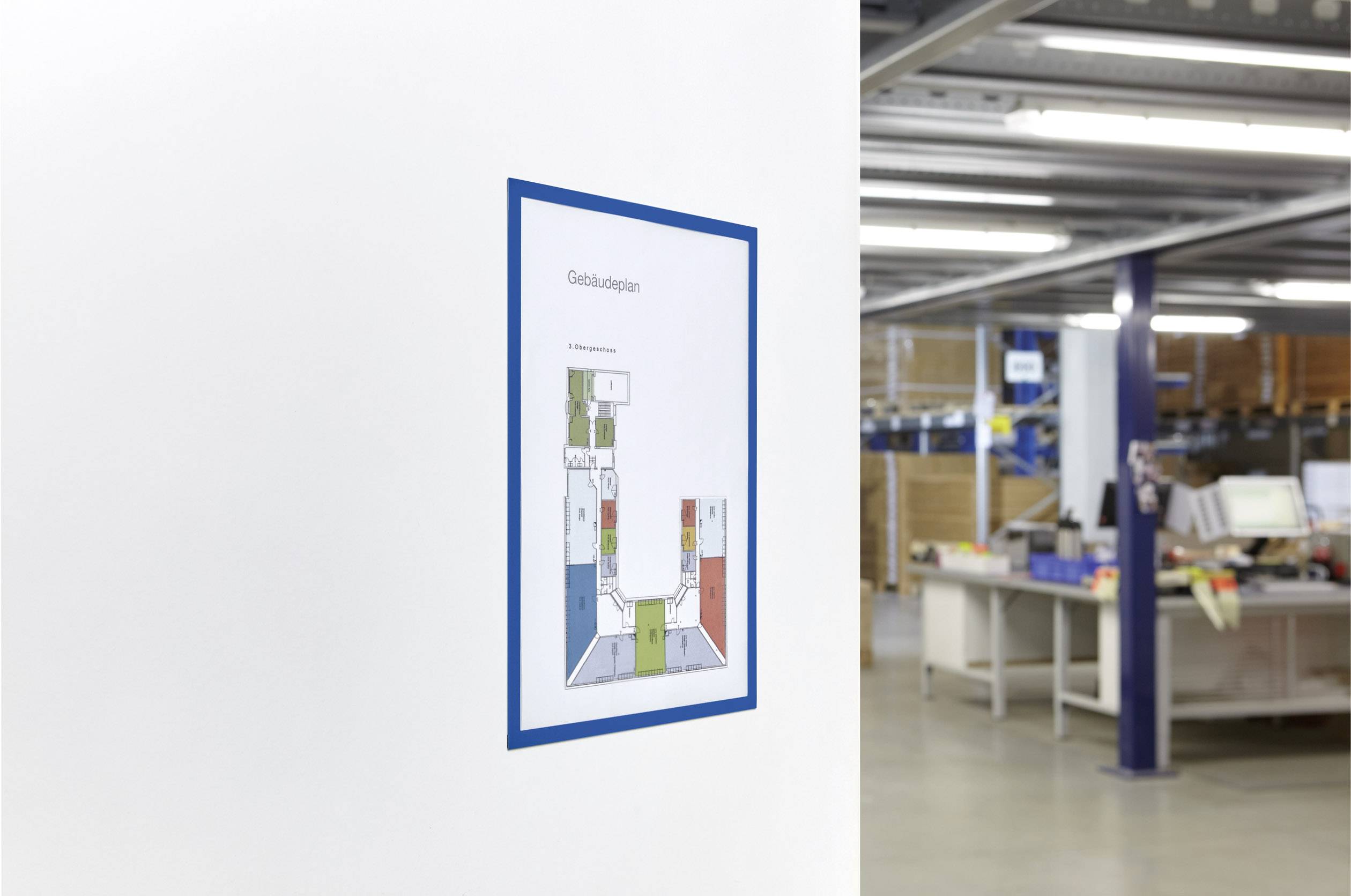 Gebäudeplan in einem blauen Rahmen, an einer weißen Wand in einer Lagerhalle hängend. Im Hintergrund sind Regale und Arbeitsplätze zu sehen.