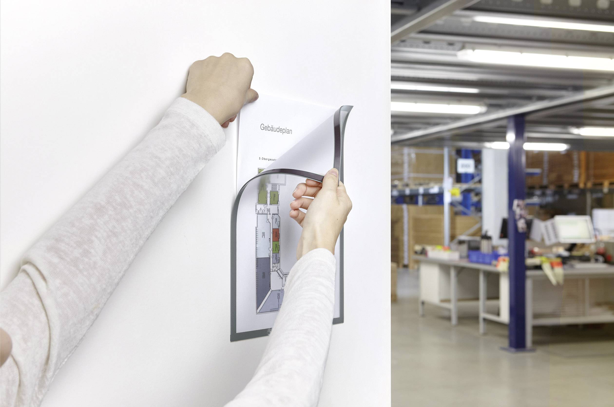 Eine Person hängt ein Gebäudeplan-Papier an eine Wand in einem Büro oder Lagerhaus auf. Im Hintergrund sind Regale und Büromaterialien sichtbar.