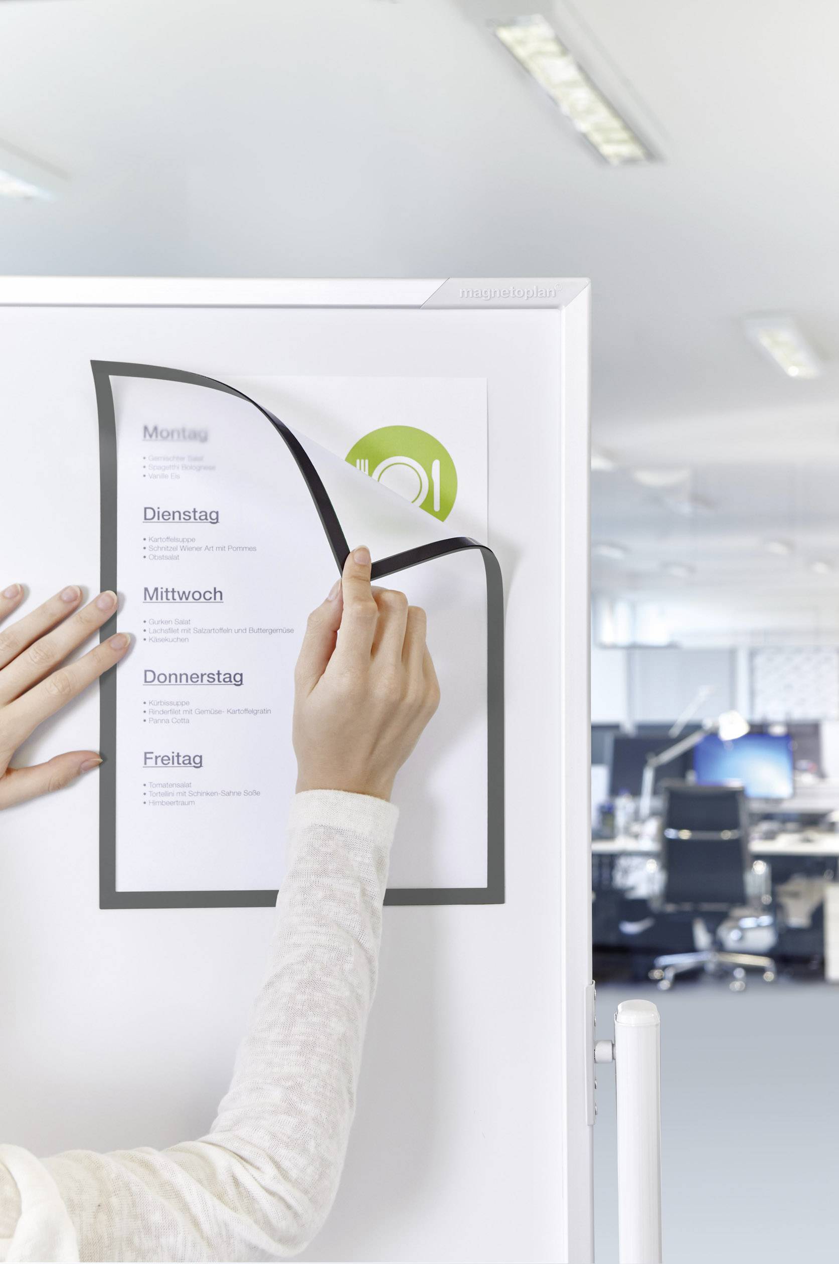'Person platziert ein Blatt Papier mit Wochentagen auf einem magnetischen Whiteboard. Büro im Hintergrund.'