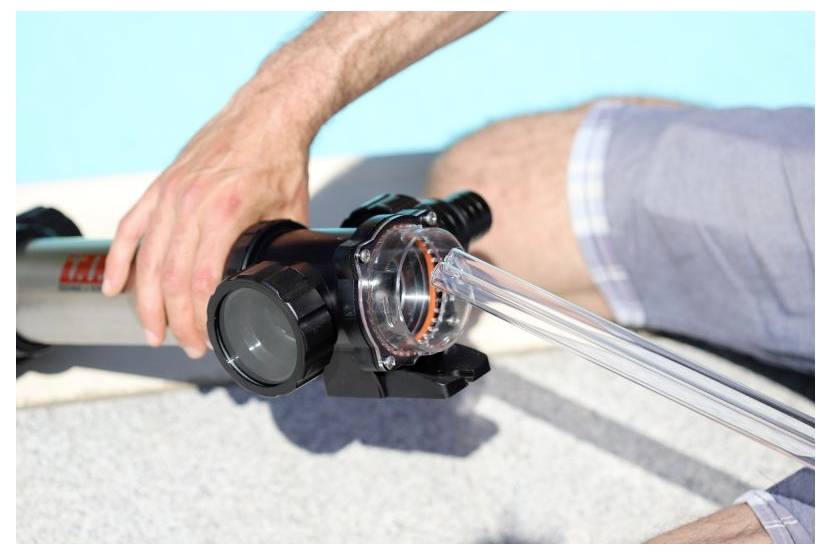 Eine Person justiert ein Poolreinigungsgerät am Rand des Wassers und konzentriert sich dabei auf die transparente Schlauchleitung, die mit einer Filtereinheit verbunden ist.