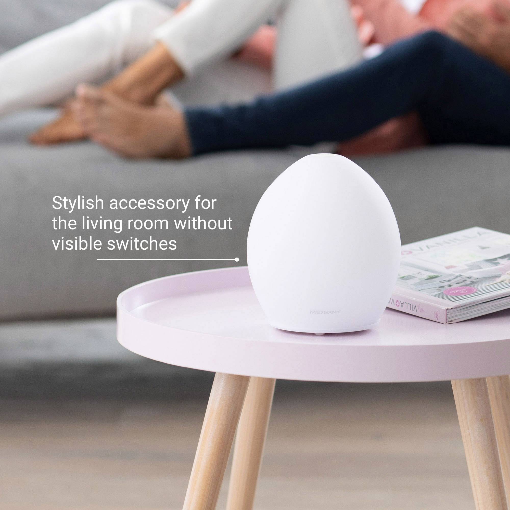 Eleganter kabelloser Lautsprecher in Ei-Form auf einem Tisch im Wohnzimmer, im Hintergrund unscharfe Personen auf einem Sofa.