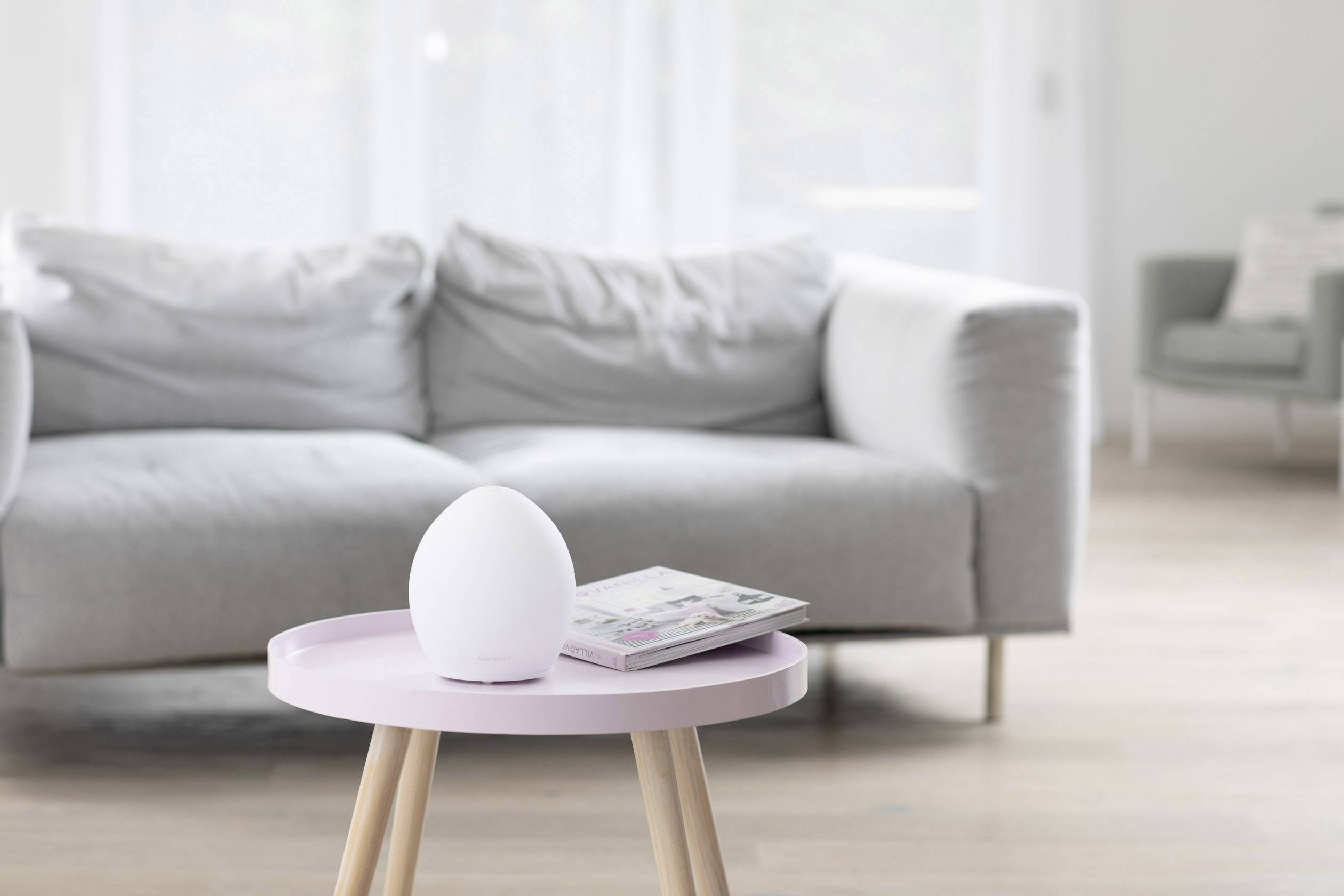 Ein Wohnzimmer mit einem grauen Sofa im Hintergrund. Im Vordergrund steht ein rosa Tisch mit einem weißen, ovalen Gerät und einer Zeitschrift.