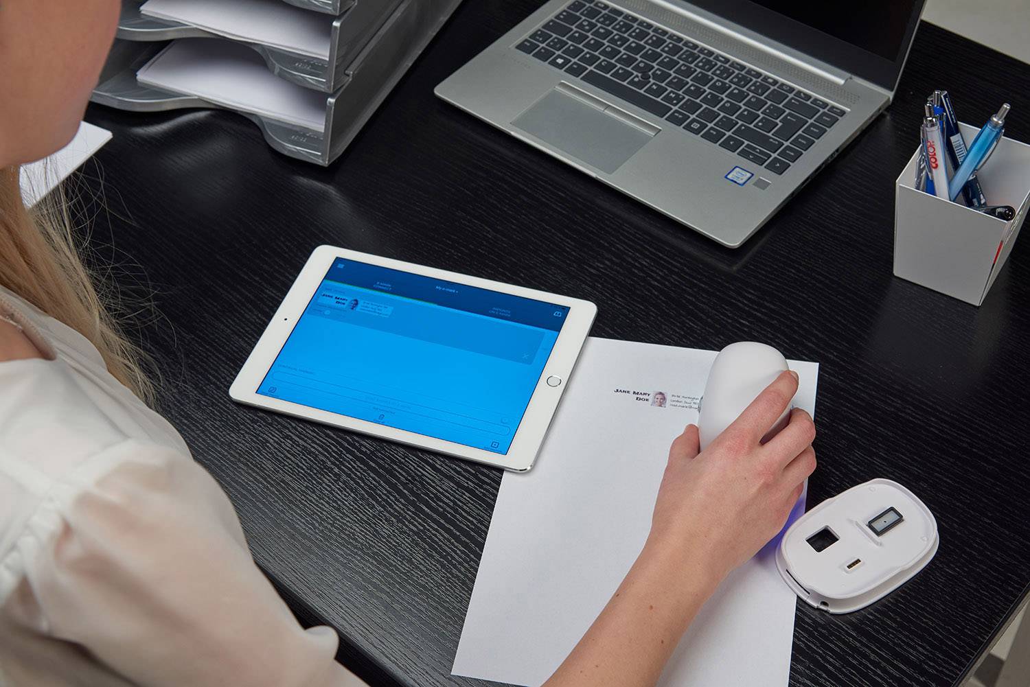 Eine Person scannt ein Dokument mit einem tragbaren Scanner. Ein Laptop und ein Tablet liegen auf einem Schreibtisch daneben.