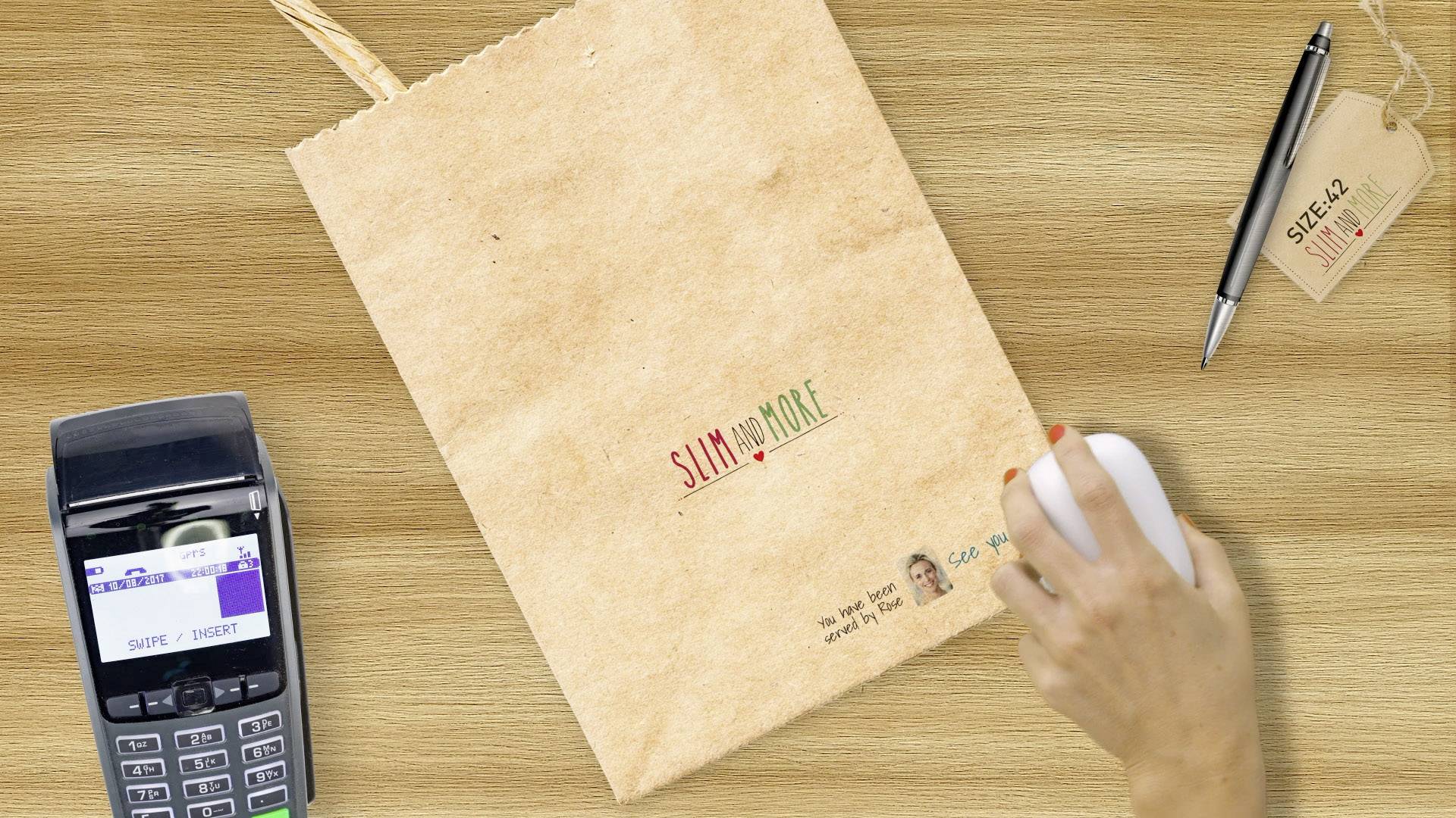 Eine Hand hält eine Computermaus neben einer braunen Papiertüte mit der Aufschrift 'Slim and More' auf einem Holztisch, daneben ein Kartenterminal.