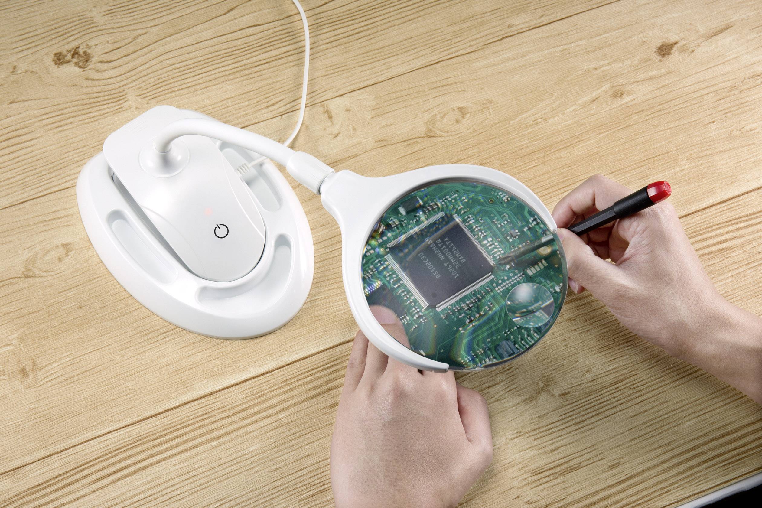 Person untersucht eine grüne Leiterplatte mit Lupe und Stift auf Holztisch. Eine elektrische Lampe bietet zusätzliche Beleuchtung.