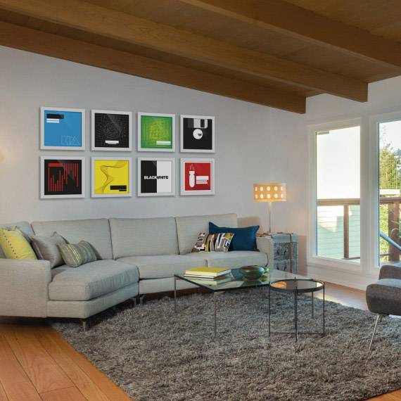 Wohnzimmer mit grauem Sofa, beigem Teppich und modernen Kunstwerken an der Wand. Ein Fenster mit Blick nach draußen.