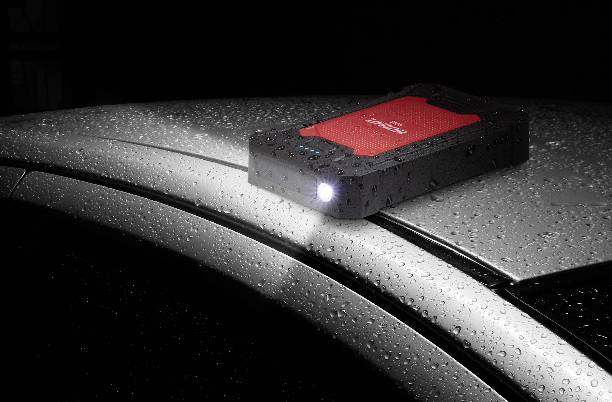 Ein rotes und schwarzes tragbares Ladegerät mit eingeschalteter Taschenlampe liegt auf einem regennassen Autodach.