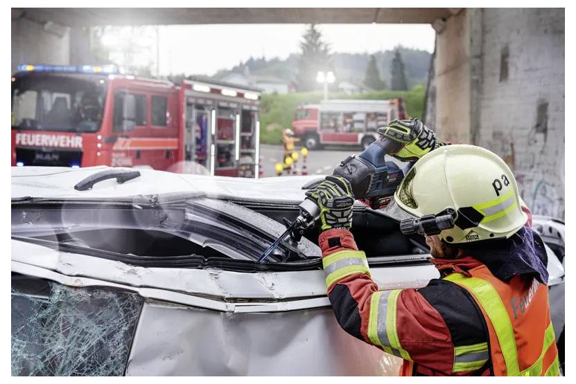Ein Feuerwehrmann in Schutzausrüstung verwendet ein Kraftwerkzeug, um das Dach eines Autos bei einer Rettungsaktion abzuschneiden, während Feuerwehrfahrzeuge im Hintergrund stehen.