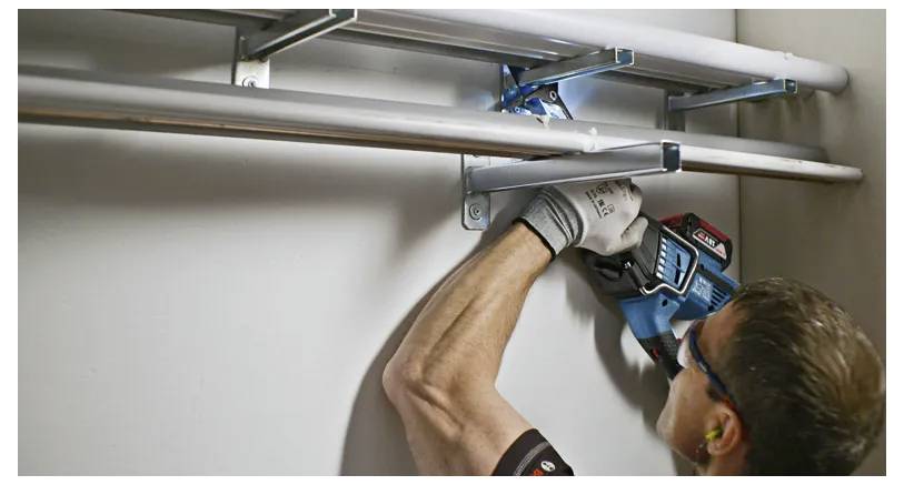 Eine Person mit Handschuhen benutzt eine Bohrmaschine, um drei Metallstangen an einer Wand zu installieren und konzentriert sich dabei auf eine Befestigungshalterung.