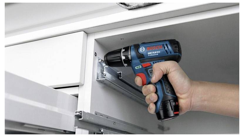 Eine Person verwendet eine blaue und schwarze kabellose Akkuschrauber, um eine Schubladenführung an einem weißen Schrank zu installieren.