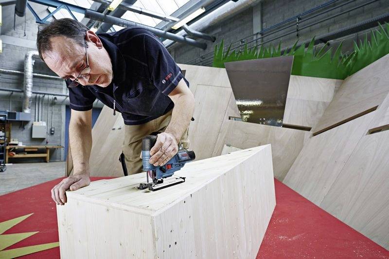 Eine Person benutzt ein Elektrowerkzeug, um eine große Holzkonstruktion in einer Werkstattumgebung zu schneiden, umgeben von kantig geformten Holzpaneelen.