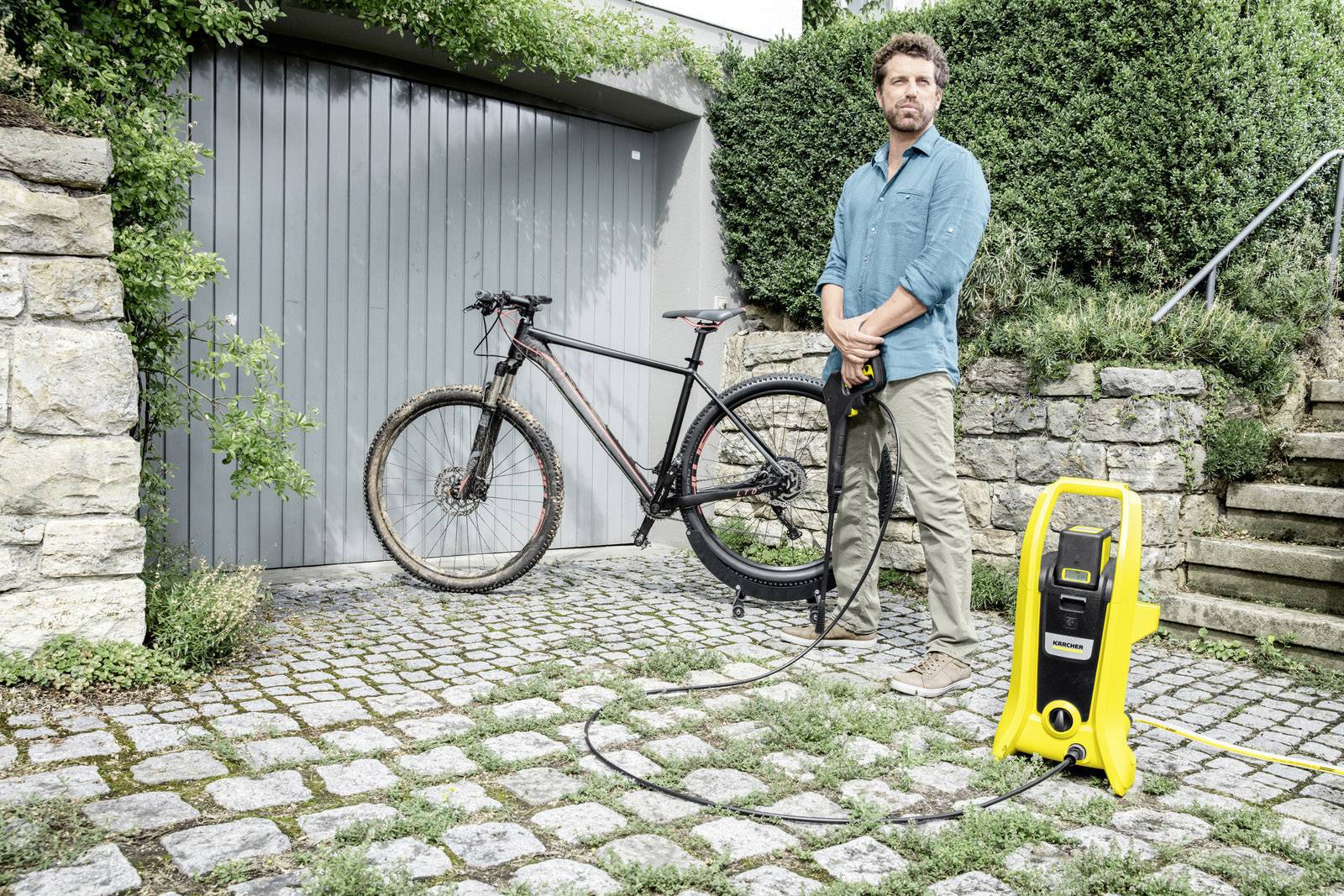 Ein Mann reinigt ein schwarzes Fahrrad mit einem Hochdruckreiniger vor einer grauen Garage auf gepflastertem Boden.