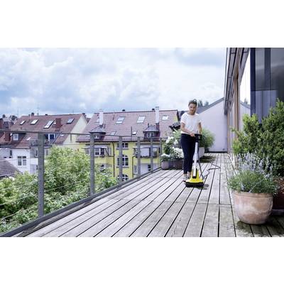 Eine Person reinigt eine Holzterrasse mit einem gelben Hochdruckreiniger. Im Hintergrund sind Wohnhäuser und Bäume zu sehen.