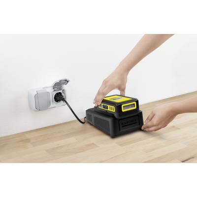 Hände, die eine gelb-schwarze Batterie in ein markiertes Ladegerät auf einer Holzoberfläche einsetzen, wobei ein Kabel in eine Wandsteckdose gesteckt ist.