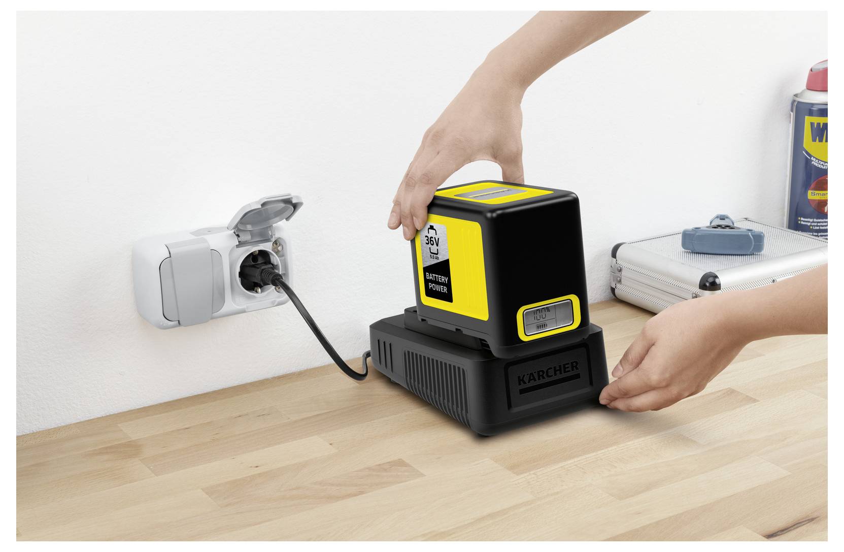 Eine Person legt eine gelb-schwarze Batterie auf ein Ladegerät auf einer Holzoberfläche. Eine Steckdose mit Stecker ist an der Wand sichtbar.