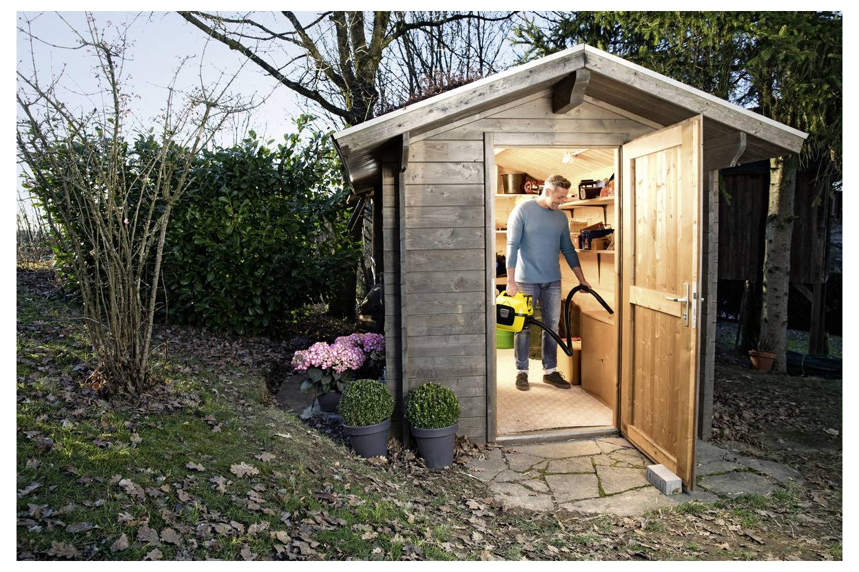Eine Person in einem Gartenschuppen, die einen Laubbläser hält, mit der Tür offen. Pflanzen und Bäume umgeben den Schuppen auf einer Rasenfläche.