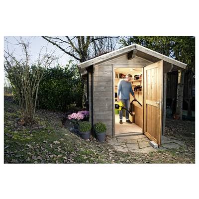 Eine Person in einem Gartenschuppen, die einen Laubbläser hält, mit der Tür offen. Pflanzen und Bäume umgeben den Schuppen auf einer Rasenfläche.