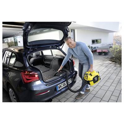 Ein Mann benutzt einen gelben tragbaren Staubsauger, um den Kofferraum eines blauen Autos zu reinigen, das auf einer Auffahrt geparkt ist.