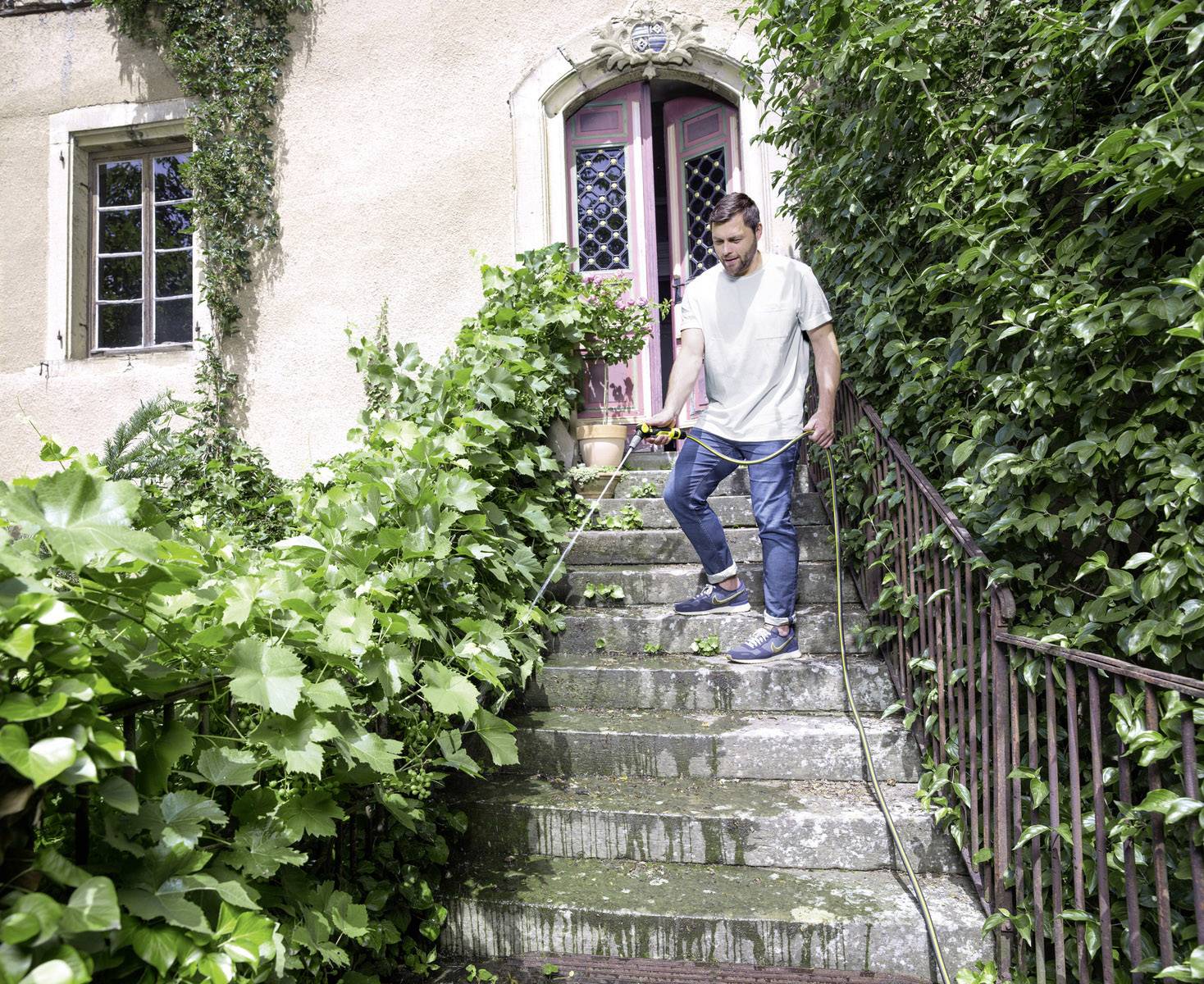 Eine Person steigt die von Pflanzen gesäumte Steintreppe eines Hauses hinunter. Im Hintergrund sind die violetten Türen des Hauses sichtbar.