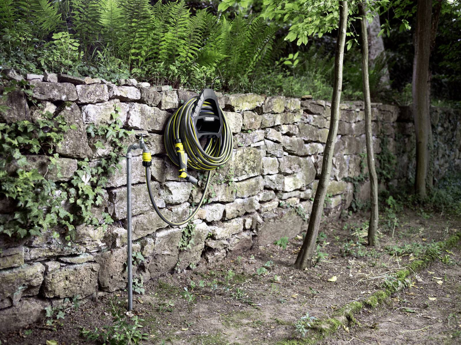 Ein Gartenschlauch hängt aufgerollt an einer Steinmauer, umgeben von grünen Pflanzen und Bäumen. Der Boden ist erdig.