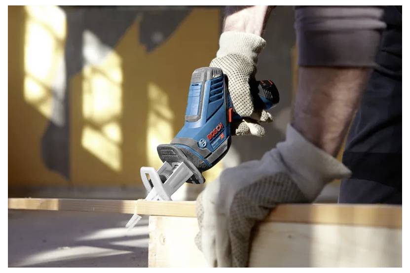 Eine Person mit Handschuhen verwendet eine blaue und schwarze Kreissäge, um ein Holzbrett im Innenraum zu schneiden, während Sonnenlicht durch die Fenster hereinströmt.