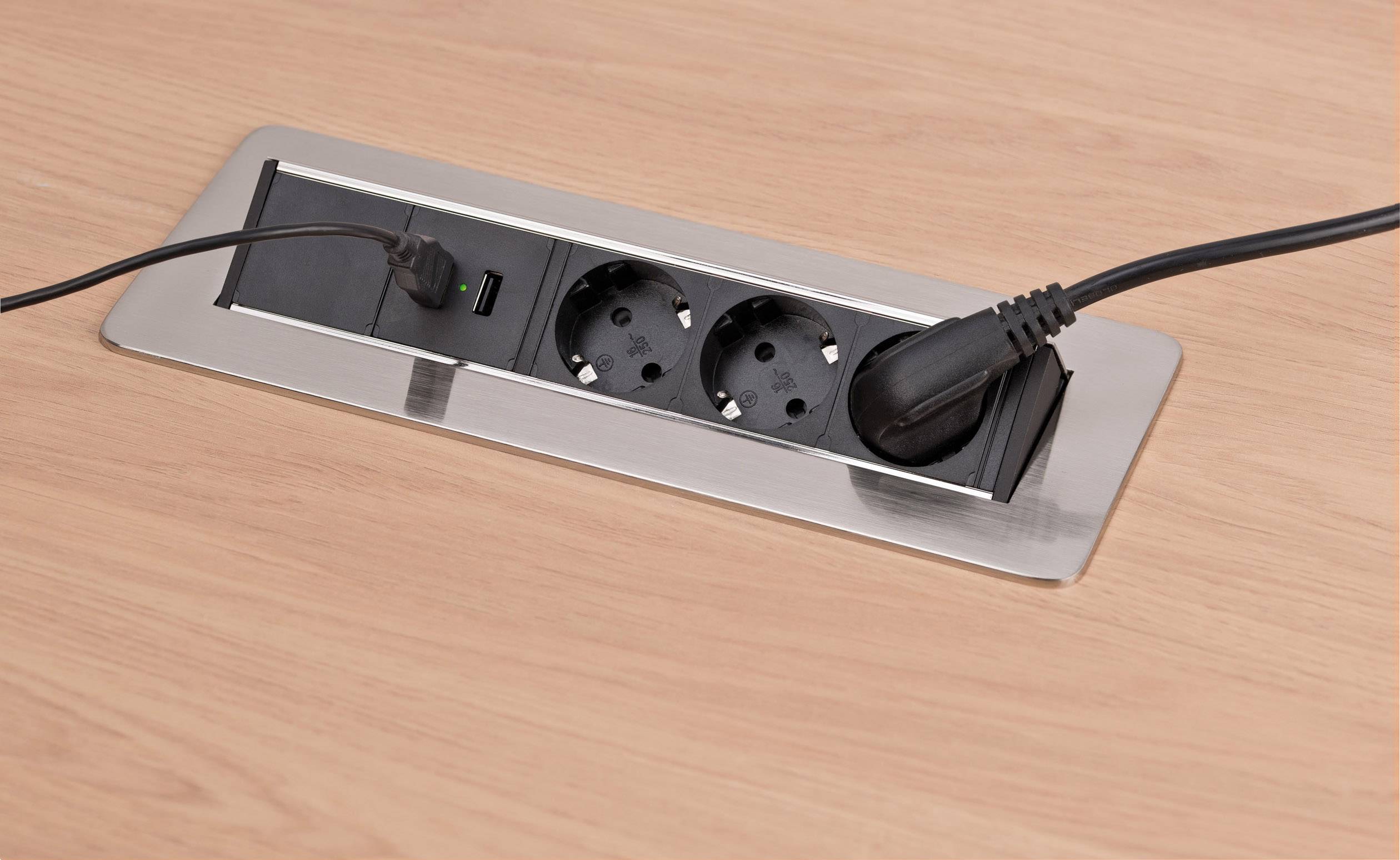 In einem Tisch integrierte Steckdosenleiste mit zwei runden Steckdosen; ein schwarzes und ein dünnes Kabel sind angeschlossen.