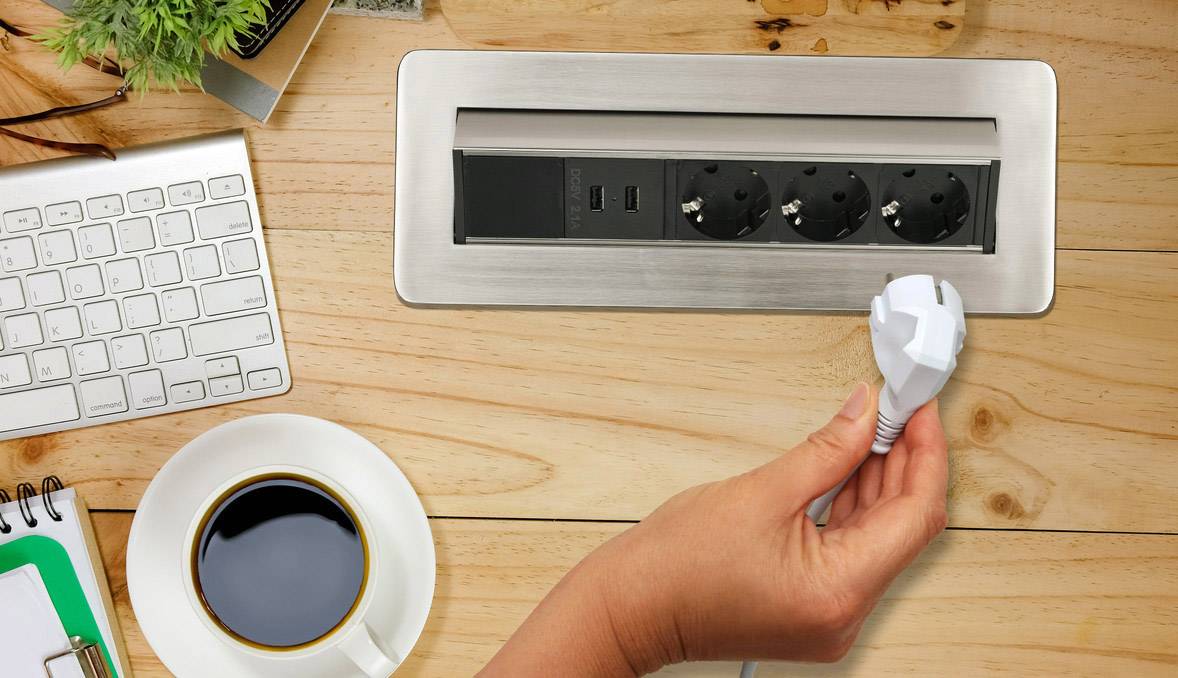 Eine Hand hält einen Stecker über einem Schreibtisch mit Tastatur, Kaffeetasse und Steckdosenleiste, bereit zum Einstecken.