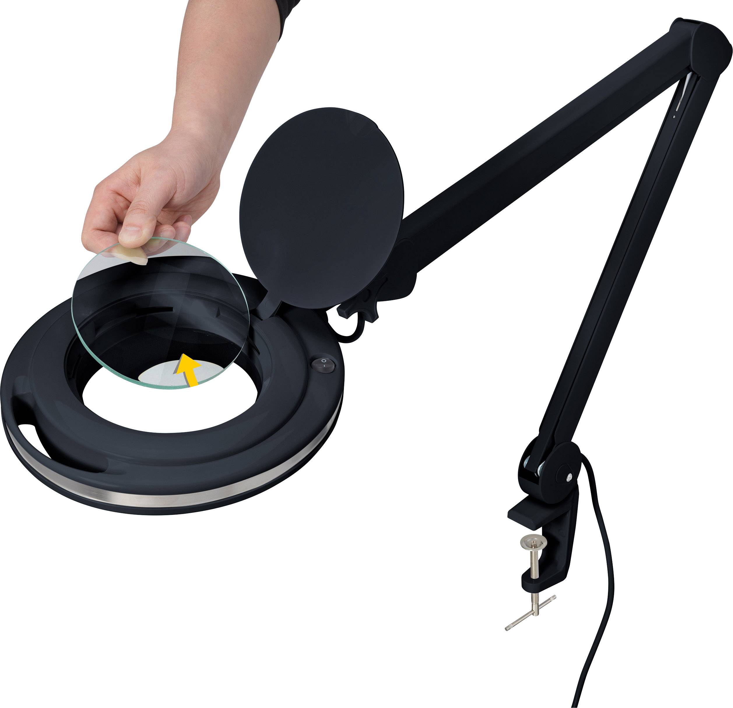 Eine Hand legt eine Linse in eine runde, schwarze Lupenlampe mit einem langen, verstellbaren Arm und einer Tischklemme.