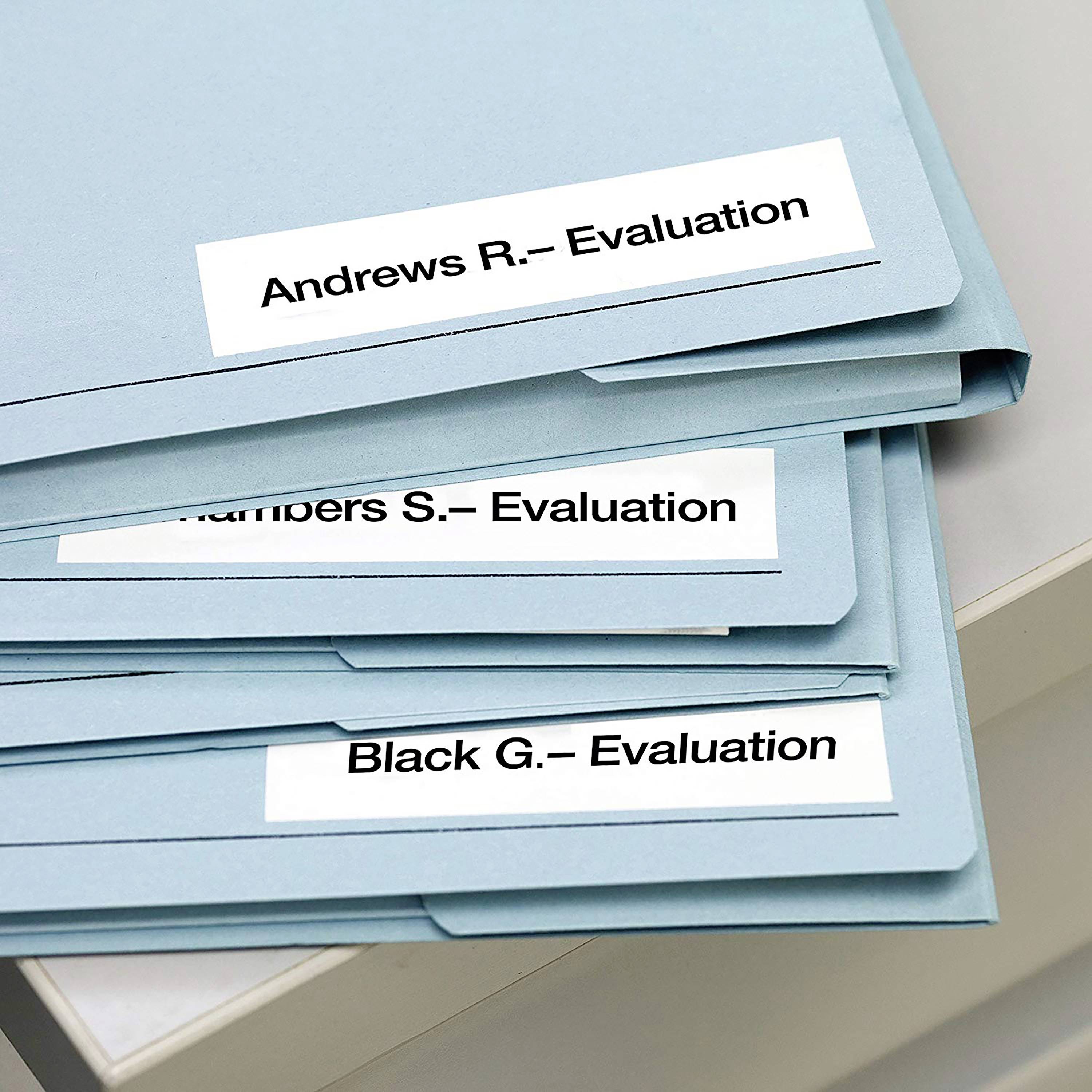 Drei blaue Aktenordner mit Beschriftungen: 'Andrews R. - Evaluation', 'Chambers J. - Evaluation', 'Black G. - Evaluation', auf einem Tisch.