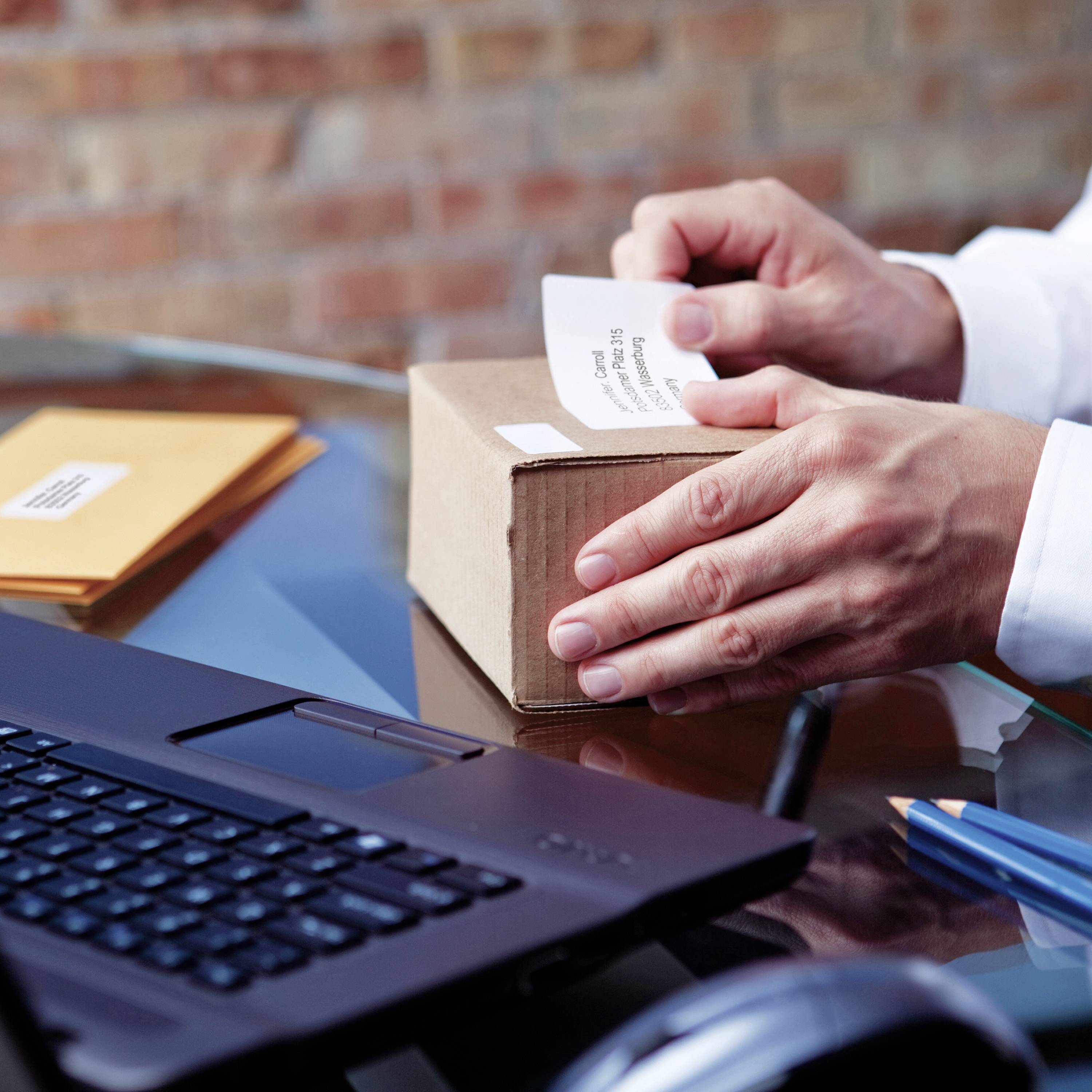 Person öffnet ein Paket auf einem Tisch, neben einem Laptop. Büroarbeitsplatz mit Versandumschlägen, ein Hinweis auf eine Arbeitssituation.