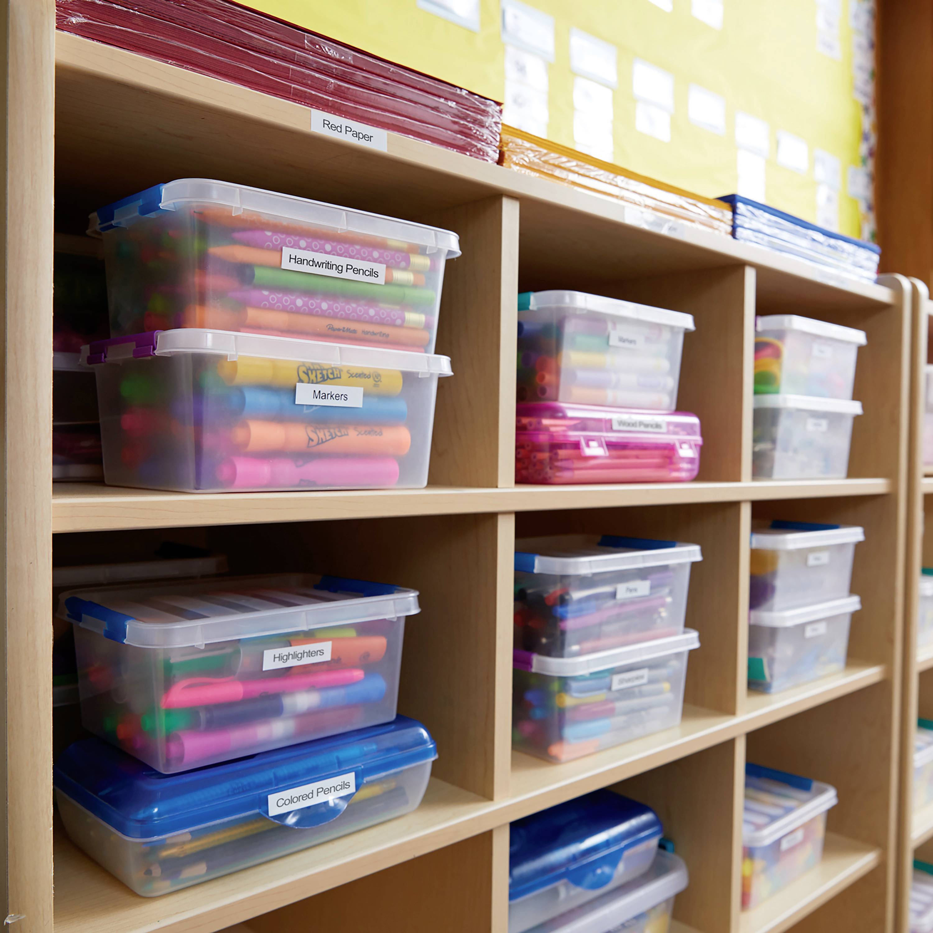 Regale mit beschrifteten Plastikboxen, die verschiedene Schreibwaren wie Marker und Stifte enthalten, in einem Klassenzimmer.