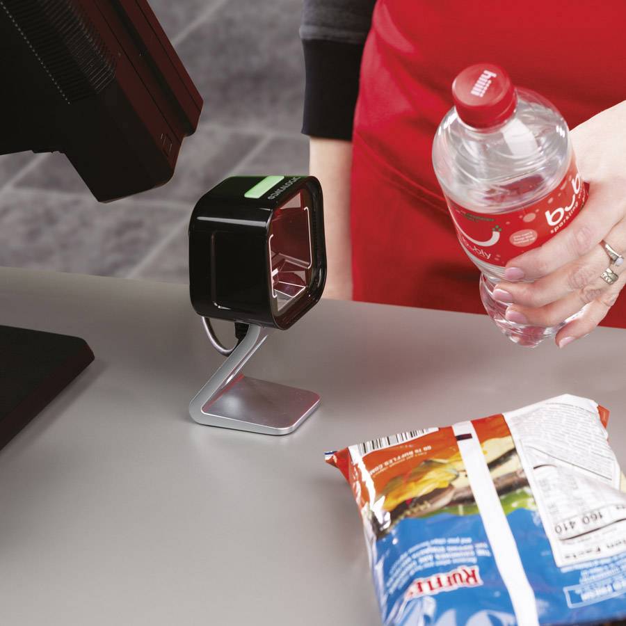 Eine Person in rotem Schürze scannt Produkte an der Kasse. Eine Flasche und eine Chipstüte liegen auf dem Tresen.