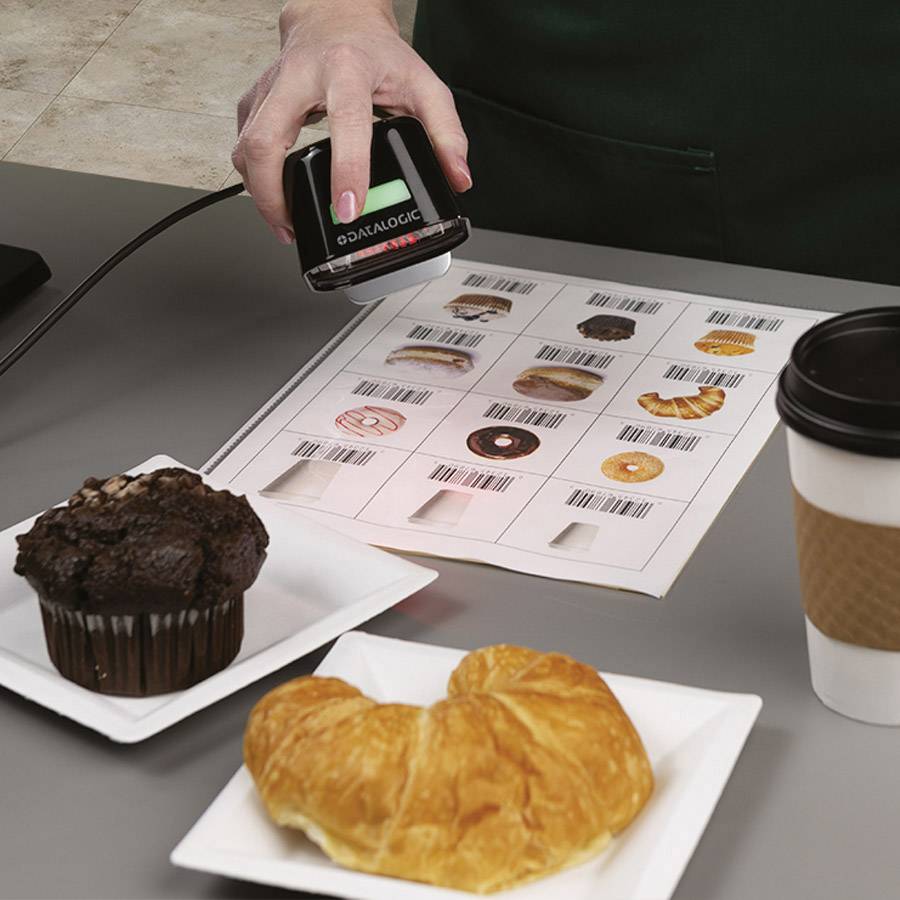 Eine Hand hält einen Barcodeleser über ein Blatt mit Barcodes, daneben stehen ein Schokoladenmuffin und ein Croissant auf Tellern.