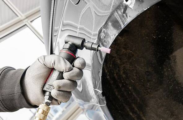 Eine Person mit Handschuhen verwendet ein Druckluftgerät zum Schleifen eines Fahrzeugkotflügels, was laufende Autoreparaturarbeiten anzeigt.