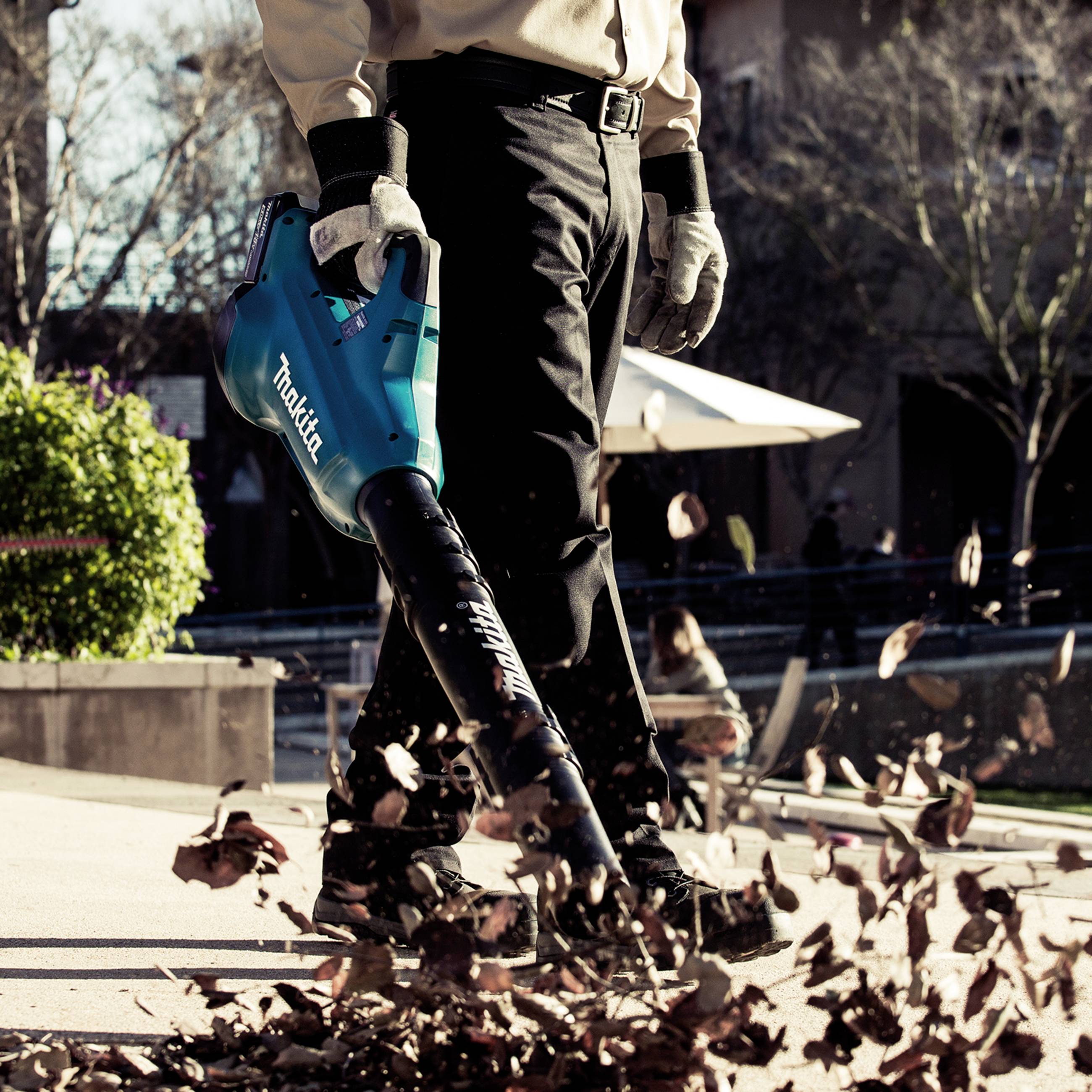 Eine Person verwendet einen Laubbläser, um Herbstblätter auf einem Bürgersteig zu entfernen. Im Hintergrund sind Bäume und Gebäude.