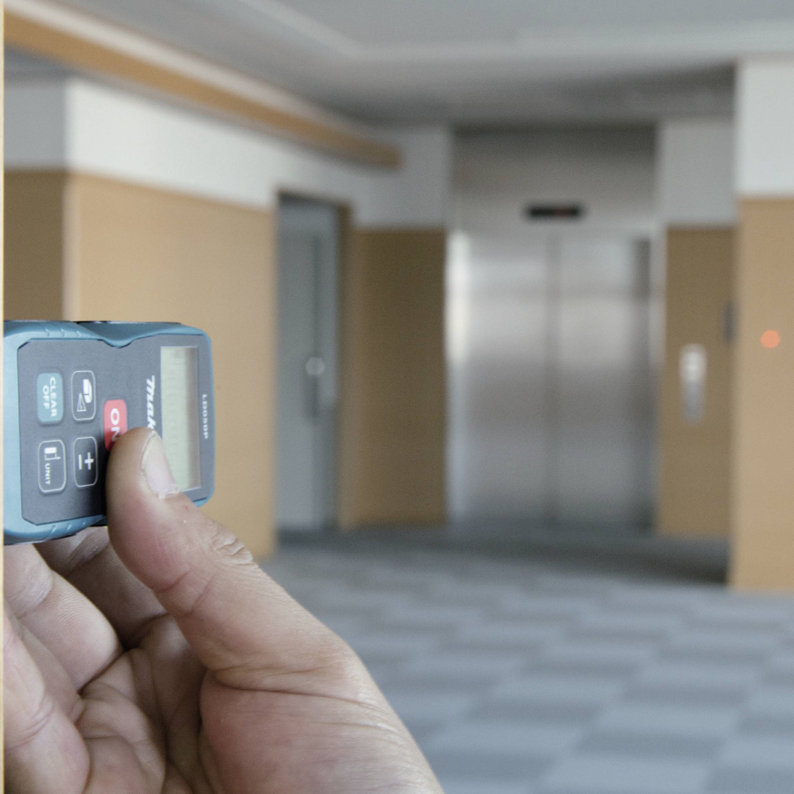 Eine Hand hält eine Fernbedienung in einem Gebäude, während im Hintergrund ein Aufzug sichtbar ist.
