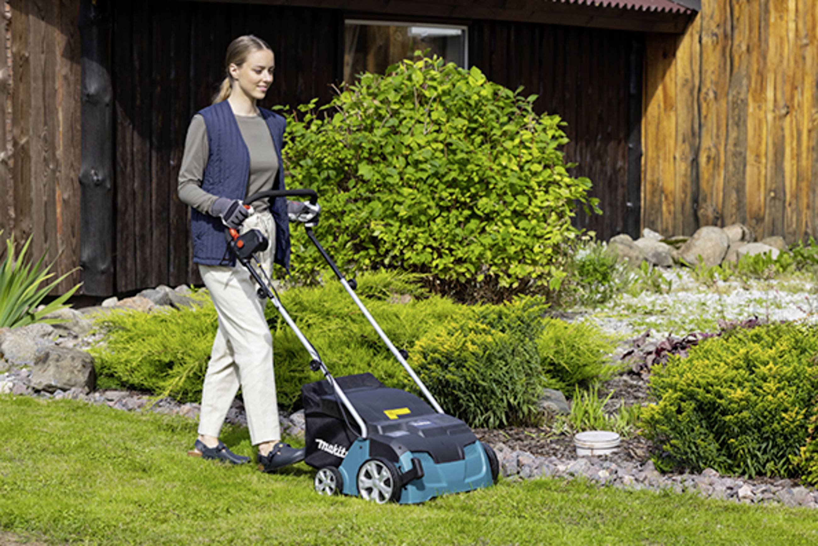 Eine Frau mäht den Rasen in einem Garten mit einem elektrischen Rasenmäher, umgeben von grünen Büschen und einem Holzhäusern.